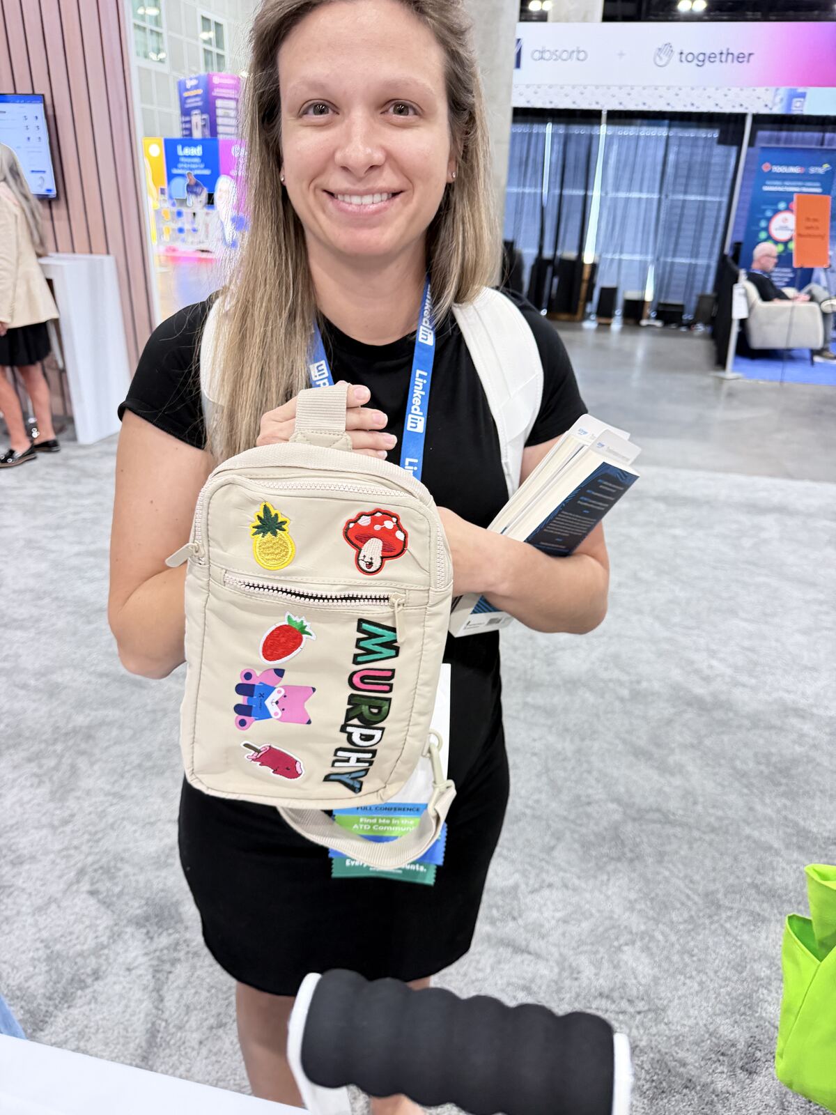 A person holds an embroidered canvas tote bag at an event booth with branded merchandise displays
