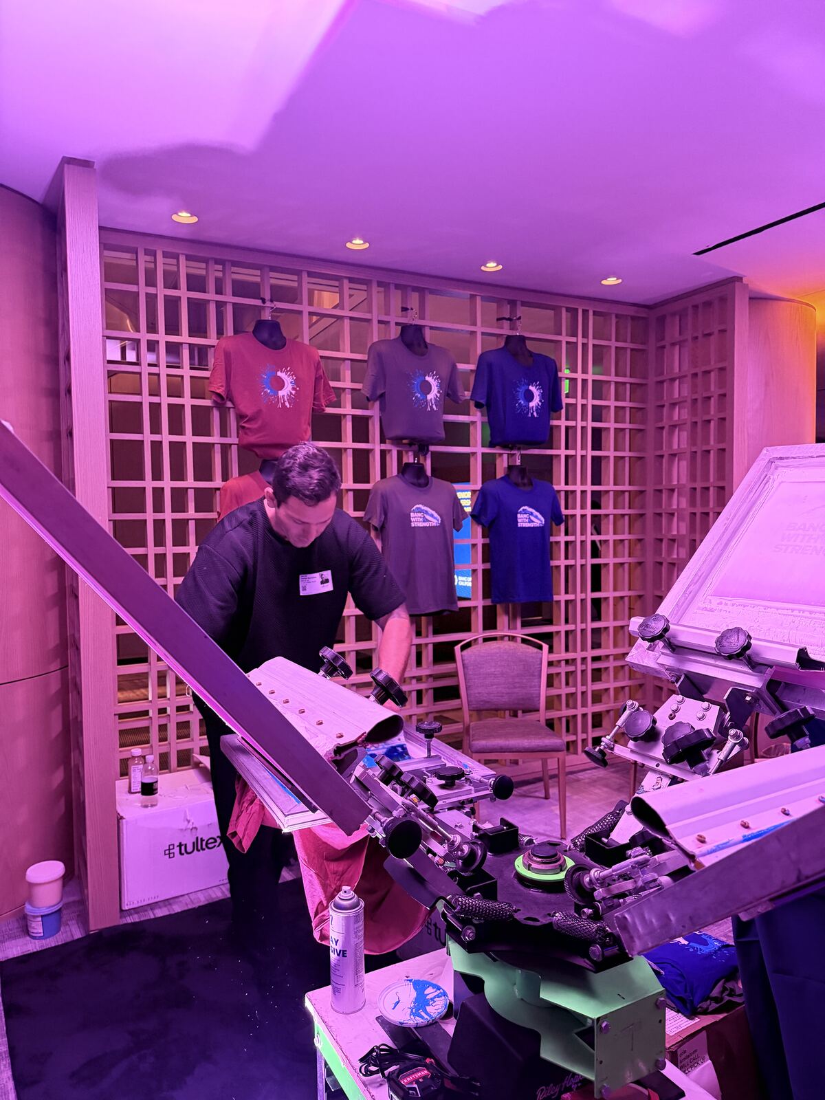 Staff member operating screen printing equipment with custom-decorated tshirts displayed on a gridded wall under ambient purple lighting